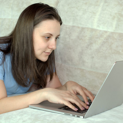 Eine Frau liegt auf einem Sofa, schaut auf einen Laptop und hat die Häde auf der Tastatur.