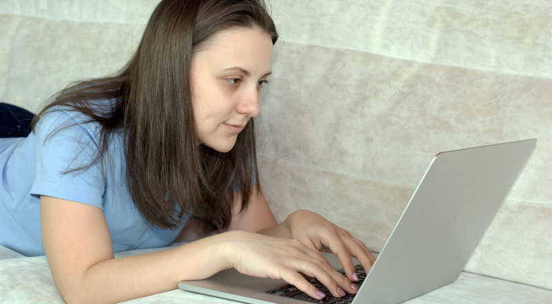 Eine Frau liegt auf einem Sofa, schaut auf einen Laptop und hat die Häde auf der Tastatur.